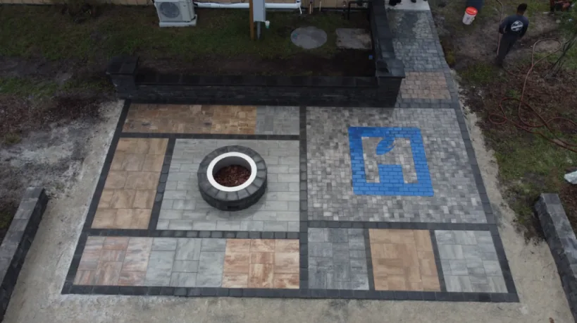 Aerial view of a geometric paver patio in gray, tan, and rust with black borders, a round fire pit, and a blue painted mark, with bare dirt and a worker at the edge in St. Johns County FL