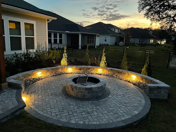 Circular paver fire pit patio at dusk with curved seat wall, landscape lighting, and illuminated cypress trees in St. Johns County FL