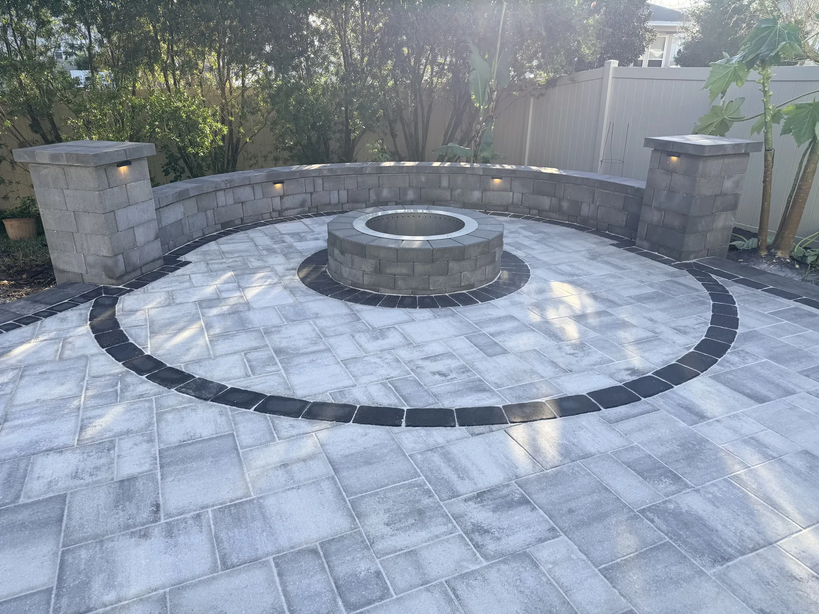 Daytime circular gray paver patio with black ring border, round fire pit with steel insert, curved seat wall, and stone pillars in St. Johns County FL