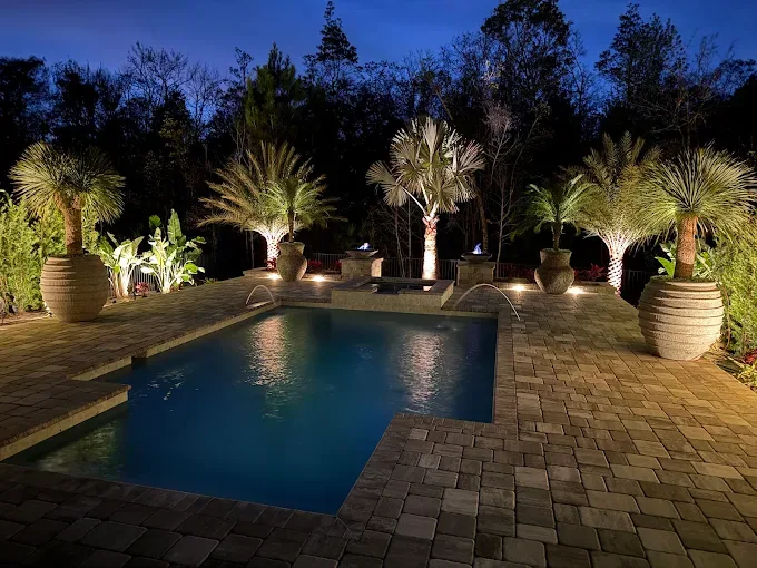 Luxury rectangular swimming pool at night with paver deck, fire bowls, and lit palm trees in decorative planters in St. Augustine FL
